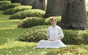 Girl dressed in white and sitting crossed logged in a beautiful green park with trees and hedges, meditating