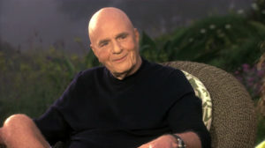 Dr Wayne Dyer sitting in a chair and smiling, bald wearing a black shirt
